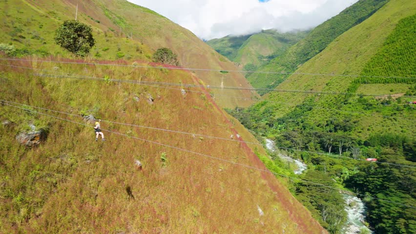 A spectacular aerial tracking shot follows a tourist who soars on a zipline high above a beautiful river valley in the Andes on the Inca Jungle Tour to Machu Picchu, Peru.
