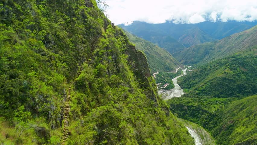 An epic aerial drone shot flies close to a steep mountainside, revealing hikers on a narrow, ancient Inca trail that overlooks a stunning green river valley on the trek to Machu Picchu, Peru.