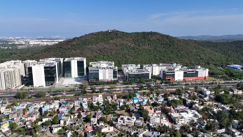 Aerial view of a large, modern IT park in Chennai at a green hill's base. It shows contrasting residential areas, a railway, and roads, highlighting urban growth and nature.