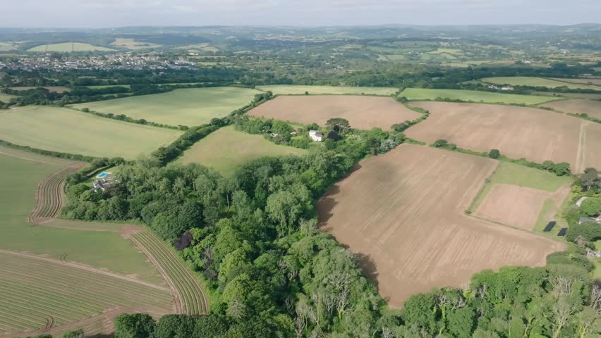 Farmhouses nestled in vibrant green woodland surrounded by farmed fields lined with hedgerows. Summer, Cornwall, UK.