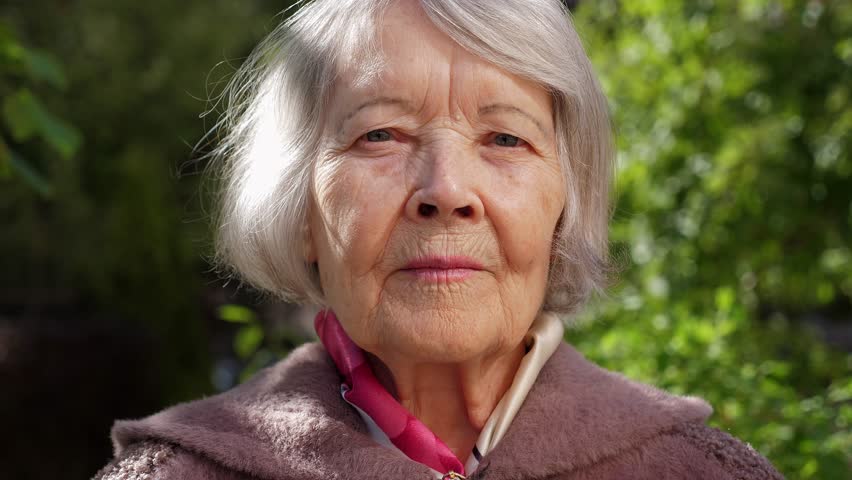 Close-up of the face of an old grandmother with gray hair in the park, she stands against the background of blurred trees and looks into the camera. There are many wrinkles on the old woman