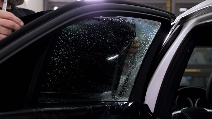 A close-up mechanic installs a tinting film on the car glass, he uses a spatula to remove excess water from the glass. The process of installing a protective film and tinting on the car glass.