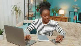 African American woman analyzing cryptocurrency data at home. Viewing charts on laptop, typing notes, checking rates on smartphone. Focused expression, working indoors, financial analysis setting. - Powered by Shutterstock - Get 15% off with code: PIKWIZARD15