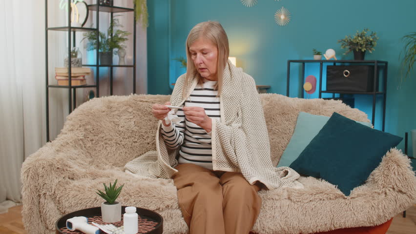 Mature woman sits on sofa covered with blanket checking temperature with thermometer coughing blows sneezes wipes snot into napkin. Grandmother shows signs of illness, including virus allergy symptoms