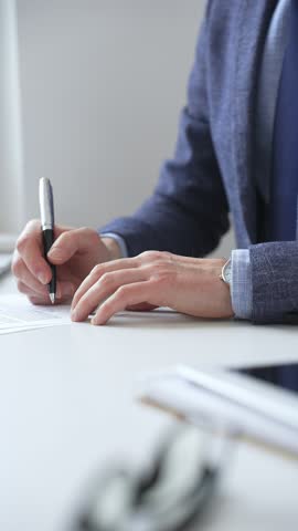 Close up of businessman's hands signing a contract with elegant pen, wearing a blue suit and a wristwatch, sitting at a white desk. Business people concept