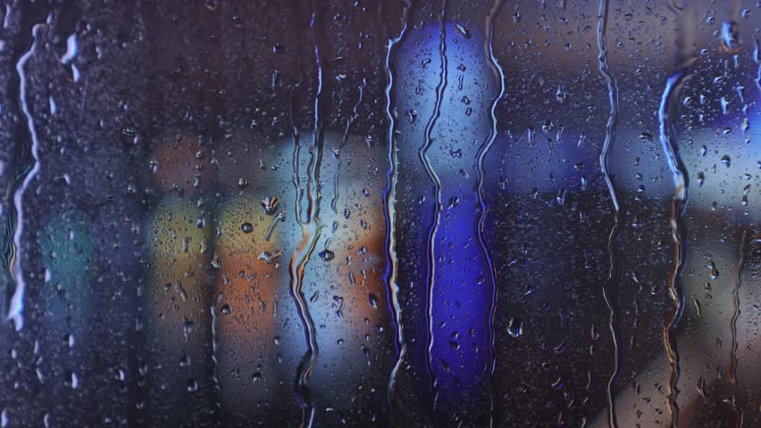 Raindrops dripping on a window pane with blurred multicolored city lights in the background at night. Bright bokeh effect creates a dreamy urban atmosphere.
