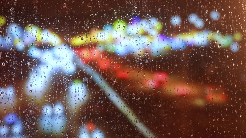 Raindrops dripping on a window pane with blurred multicolored city lights in the background at night. Bright bokeh effect creates a dreamy urban atmosphere.