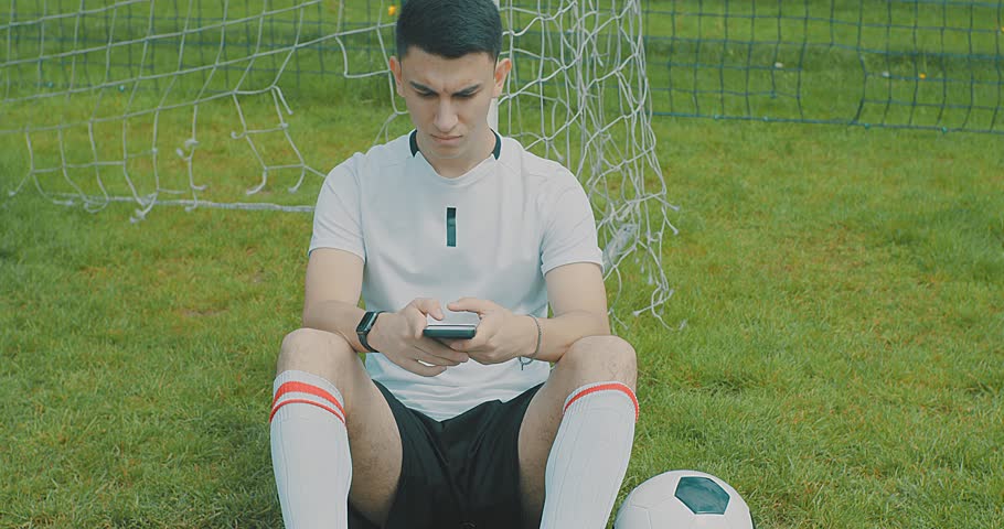 Soccer Player Sitting on Grass Near Goalpost Using Smartphone. Young soccer player resting on the grass near a goalpost while using a smartphone. After training or match, sports lifestyle, technology.