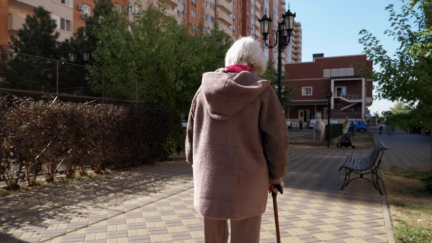 A view from the back of an old grandmother with gray hair who walks with a cane along an alley in the park. Slow motion. An old grandmother walks slowly and limps in the city.