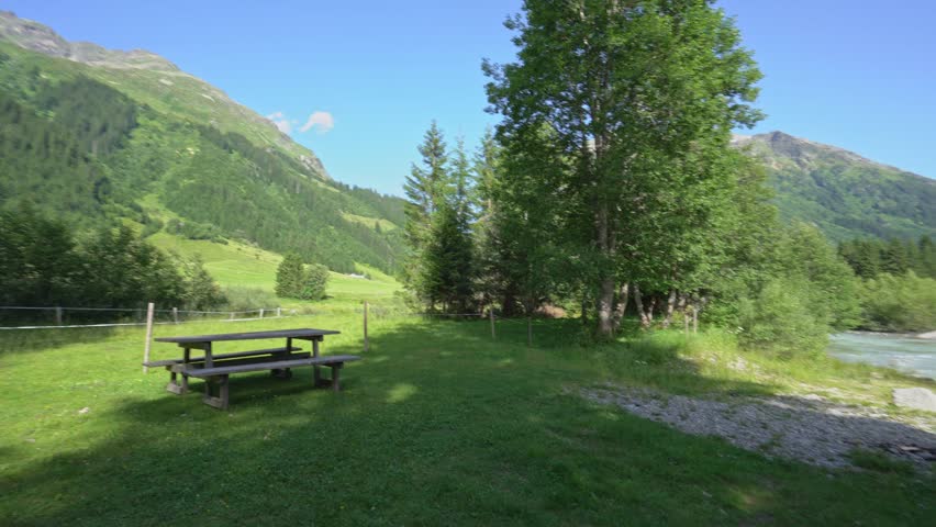 Lush green meadow with picnic table next to a clear river flowing through alpine valley