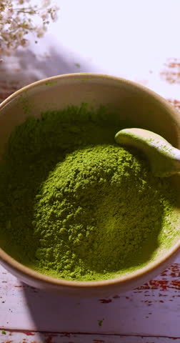 A spoon scoops up vibrant green powder and lets it fall back into the bowl in slow motion. Likely matcha, spirulina, chlorella, moringa, wheatgrass, or barley grass powder under natural daylight.