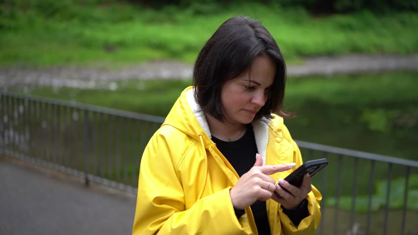 Close-up of woman in raincoat checking phone among trees. Concept: staying connected, outdoor communication, mindful use of technology.
