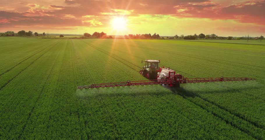 Aerial view of beautiful landscape with a crop sprayer applying pesticide in a wheat field at sunset, in the background cars and trucks passing by. Drone shot flying over agricultural wheat field