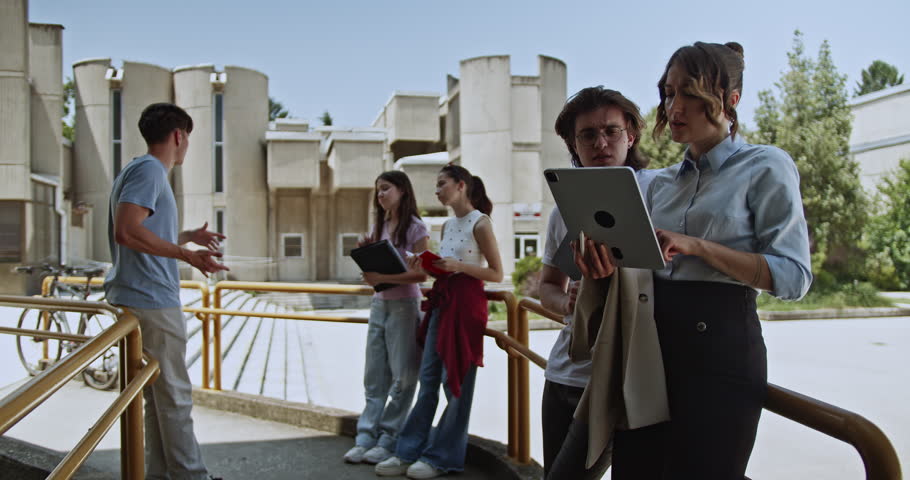 Young students and a female teacher stand on a university campus, actively engaged in a discussion. The teacher utilizes a digital tablet to facilitate their outdoor learning experience.