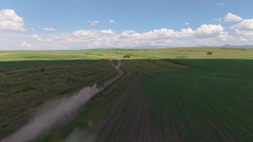 Black off road vehicle navigating dusty countryside road, leaving pronounced dust trail through verdant fields with mountain range in distant blue sky backdrop