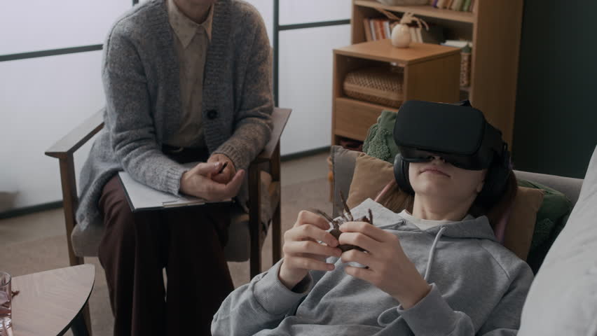 Medium shot of teenage patient lying on couch in VR headset touching fake spider to overcome arachnophobia during individual therapy session with unrecognizable psychologist