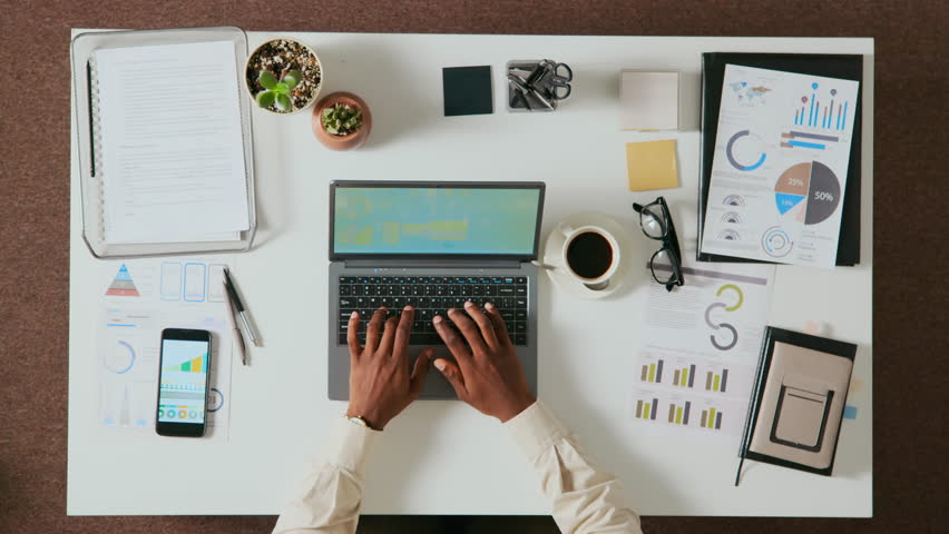 View from above of hands of unrecognisable Black man typing on laptop at white office table with smartphone, documents, post-it notes, glasses, stationery holder and cup of coffee on it