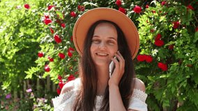 A smiling woman in a hat talks on her smartphone, with vibrant red flowers and green foliage blurring in the background. - Powered by Shutterstock - Get 15% off with code: PIKWIZARD15