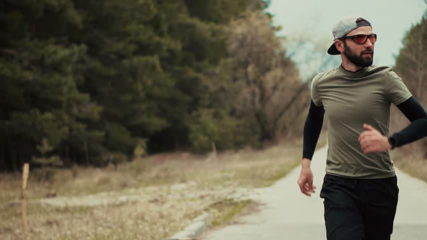 Athletic man jogging through a forest trail in cool weather, wearing sport sunglasses, cap, and layered fitness clothes. Concept of healthy lifestyle, endurance training, and outdoor running