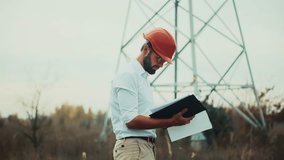 inspecting electricity object. Engineer in orange hard hat reviews documents outdoors, standing near large power pylon. energy infrastructure, professional inspection, construction engineering attire - Powered by Shutterstock - Get 15% off with code: PIKWIZARD15