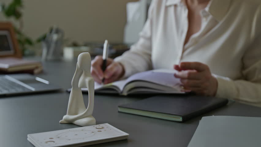 Rack focus on focused middle aged female psychiatrist with glasses writing in notebook planning therapy session schedule then typing on laptop while working at office desk