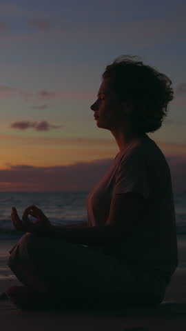 Silhouette of woman meditating in lotus pose by ocean at sunset. Camera circles around her as red and purple clouds glow in sky. Scene of peace, mindfulness and connection with nature in golden hour