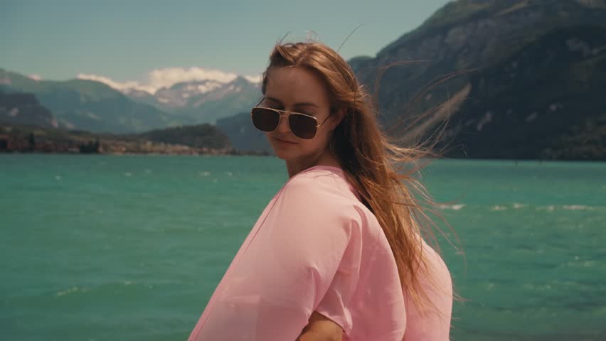 beautiful woman long blonde hair in white swimsuit pink pareo cape on standing on shore. summer background of blue sky lake mountains in Switzerland. slow motion wind blowing. Turquoise Azure Water
