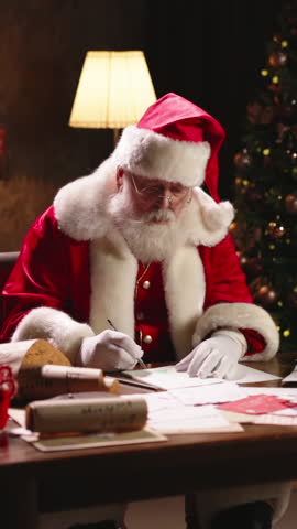 Santa Claus sitting at wooden table in cozy room, writing list of children for Christmas gifts on paper. Warm glowing lights of decorated Christmas tree and floor lamp create magical festive