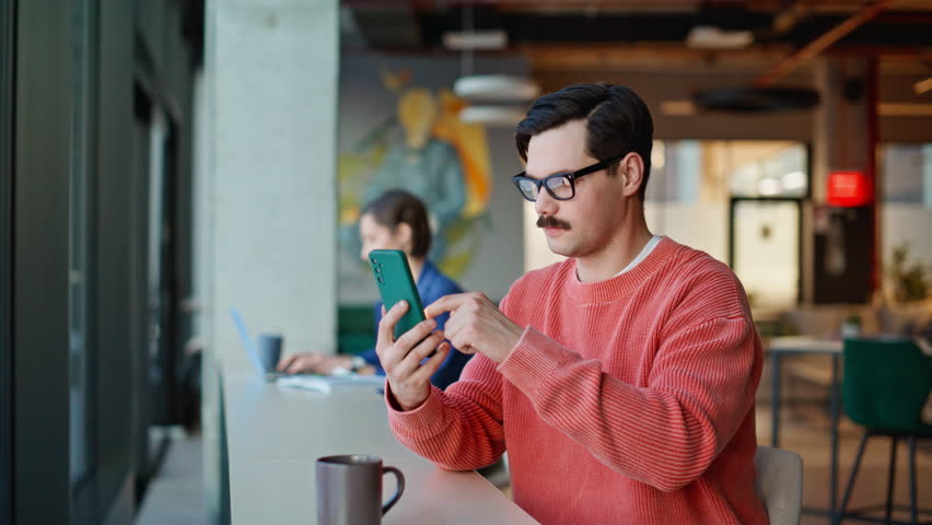 Man scrolling cellphone sitting urban cafeteria closeup. Mustached guy wearing glasses choosing online purchases using smartphone application. Dark-haired freelancer browsing mobile phone message.