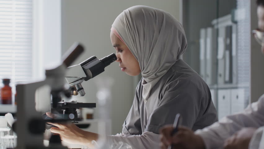 Female scientist in hijab observing slide through microscope while sitting beside male colleague taking notes at desk during research in laboratory