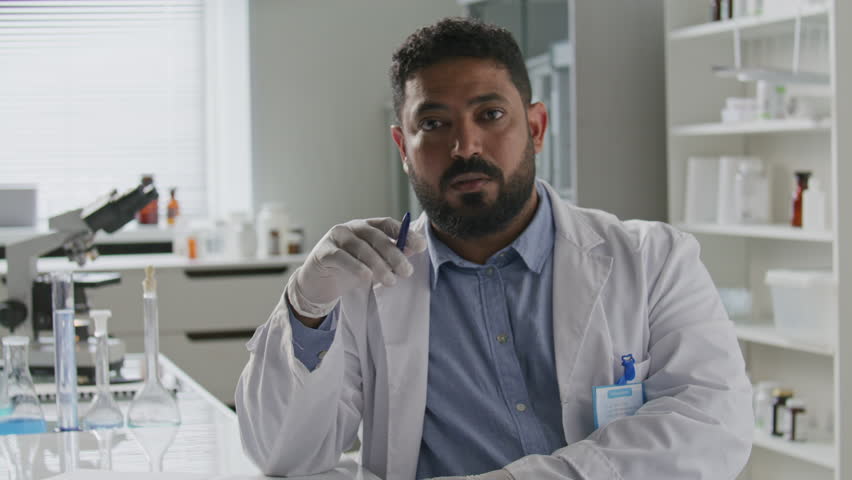 Middle eastern male scientist in gloves and lab coat looking at camera and explaining ideas during video call while sitting at desk with microscope and glassware in laboratory