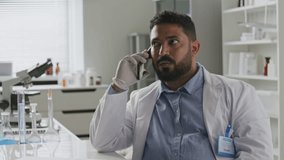 Middle eastern male scientist in lab coat and gloves speaking on mobile phone at desk with microscope and chemical glassware nearby in laboratory - Powered by Shutterstock - Get 15% off with code: PIKWIZARD15