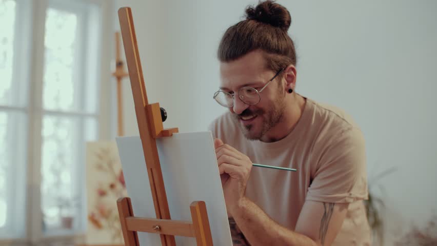 Male artist concentrating on his artwork, carefully applying paint to a canvas in his bright studio