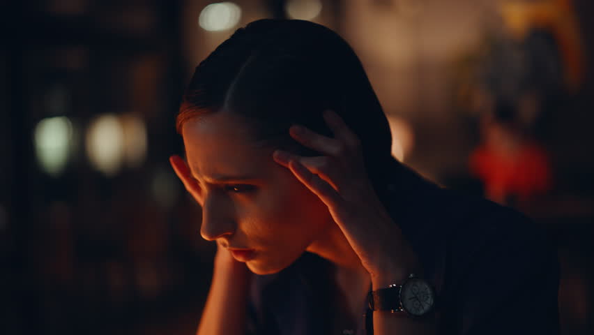 Closeup fatigued night worker touching temples finishing hard workday. Portrait tired businesswoman suffering headache of overtime work. Tensed woman manager overloaded deadline job in dark workplace.