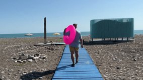 Horizontal travel video. Back view. Dolly zoom effect. White fat man in yellow shorts, blue T-shirt, flip-flops and pink circle walks along wooden path to shore on Black Sea pebble beach. Tourism - Powered by Shutterstock - Get 15% off with code: PIKWIZARD15