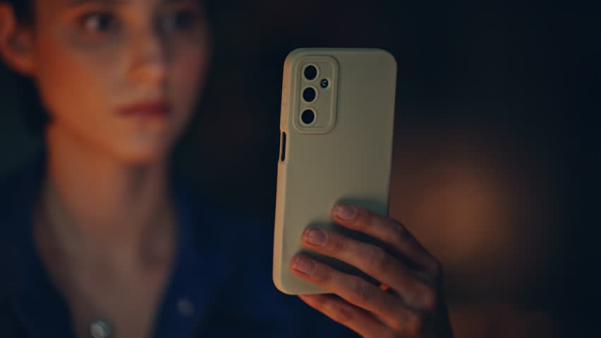 Girl watching social media at smartphone late evening closeup. Serious businesswoman reading mobile phone message at night office. Woman hand holding cellphone in dark apartment. Brunette browsing app