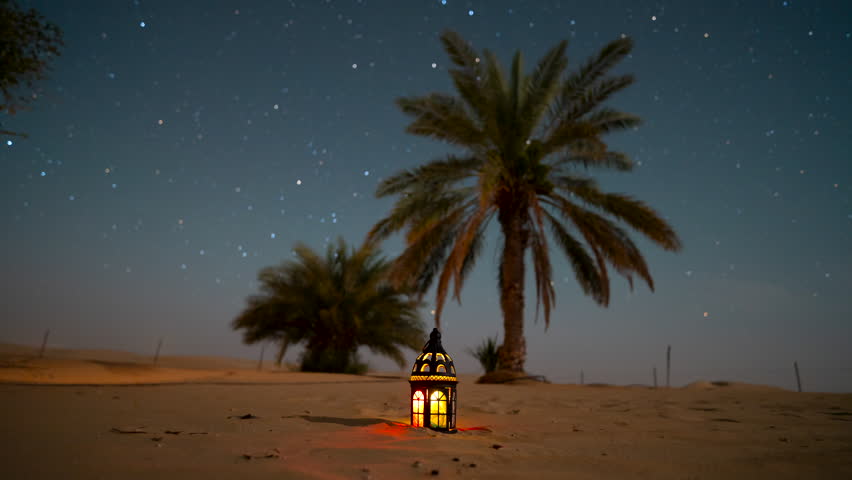 Stunning 4k time lapse of night  star sky in the desert sand dune date tree oasis with glowing Arabic lantern during Ramadan, Eid. Clear night sky in a remote wilderness location. 