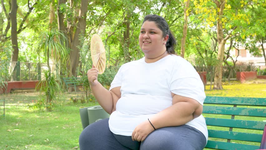 Plus size indian woman using hand fan in hot summer day. Overweight lady suffering from heat wave cooling her body in high temperature weather in india.