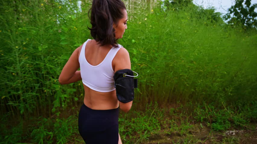 Fit girl jogs beside green meadow wearing earphones, Female Jogger in Sports Bra happily Explores Retreat, Sporty Lady Uses Armband on Trail, Lush Landscape Embraces Outdoor Runner, Health conscious