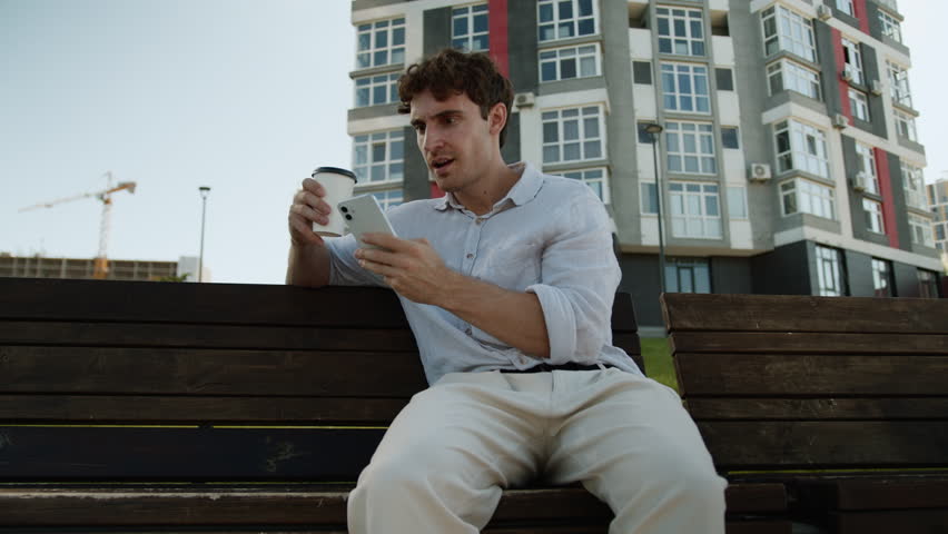 Caucasian man businessman city urban drinking coffee read news amazed male shocked mobile phone smartphone guy emotions social networks confused message stunned outdoor sitting bench business internet