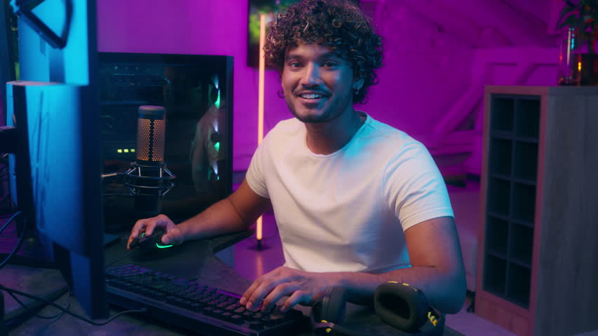 Indian Arabian Latino man playing computer pc technology game cyberspace tournament esport championship smile virtual monitor player networking online typing fun pleasure looking at camera male guy