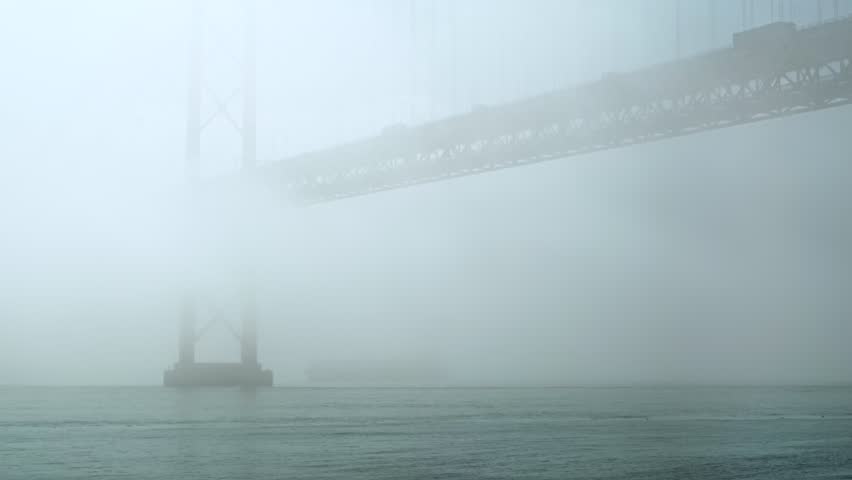 Dramatic static shot of iconic April 25th bridge in thick fog over Tagus River, Lisbon, Portugal. Beautiful view of suspension bridge with thick fog rolling over it, muted tones, traffic on bridge