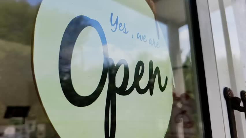 Open for Business Sign: A close-up shot of a cheerful "Open" sign displayed on a storefront window, announcing a business is welcoming customers. The sign is circular.