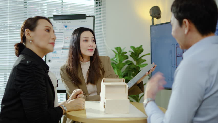 Real estate agent shows model house to two customers, discussing property details, reviewing in tablet, considering home purchase options during consultation in office, finance and investment	