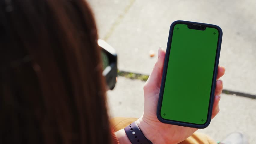 A woman sitting on bench on street background, using phone, chatting with friends on social networks, reads a book, watches a movie. Green screen mock up concept. High quality 4k footage