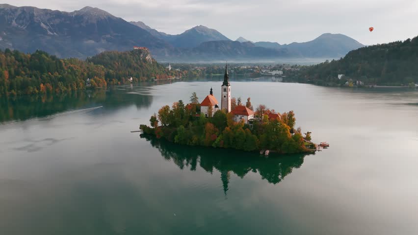 Aerial view of majestic Lake Bled at sunrise in autumn, Slovenia. Close-up of the Pilgrimage Church on the island, Bled Castle on a cliff, and a hot air balloon floating above the picturesque lake.
