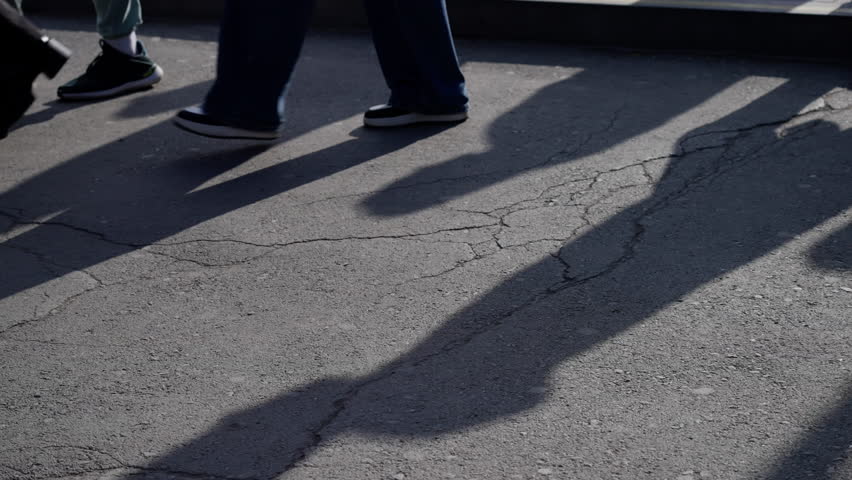 A sequence showing the movement of people walking across a sunlit, cracked pavement, casting distinct elongated shadows that slowly disappear from the frame.
