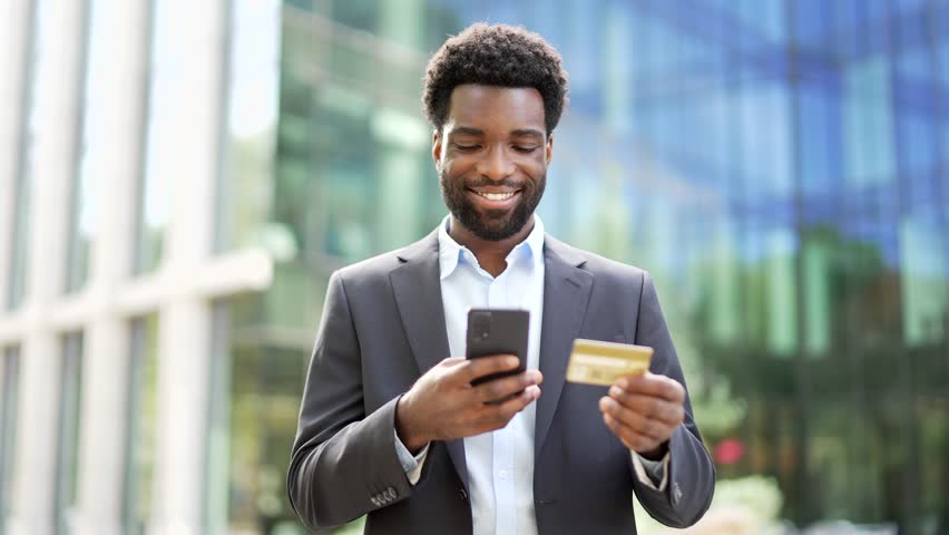 Smiling african american businessman is shopping online using credit card and mobile phone standing on street near business office building. Entrepreneur in suit happy about successful transaction