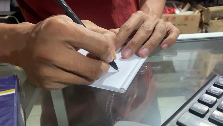 Cashier hand writing receipt on small notepad behind counter. Close-up of manual transaction process at store with visible calculator and glass counter