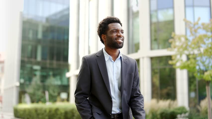 African american businessman walks down the street near an office building. Entrepreneur in formal suit walking in central business district of modern city. Handsome man worker goes to work. Close up